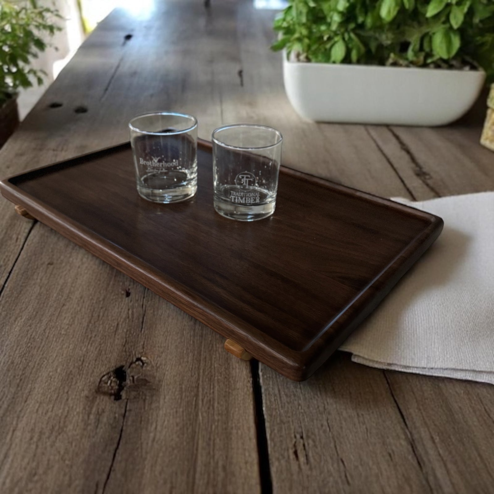 Walnut wood serving tray with maple feet sitting on an oak table with 2 glasses.